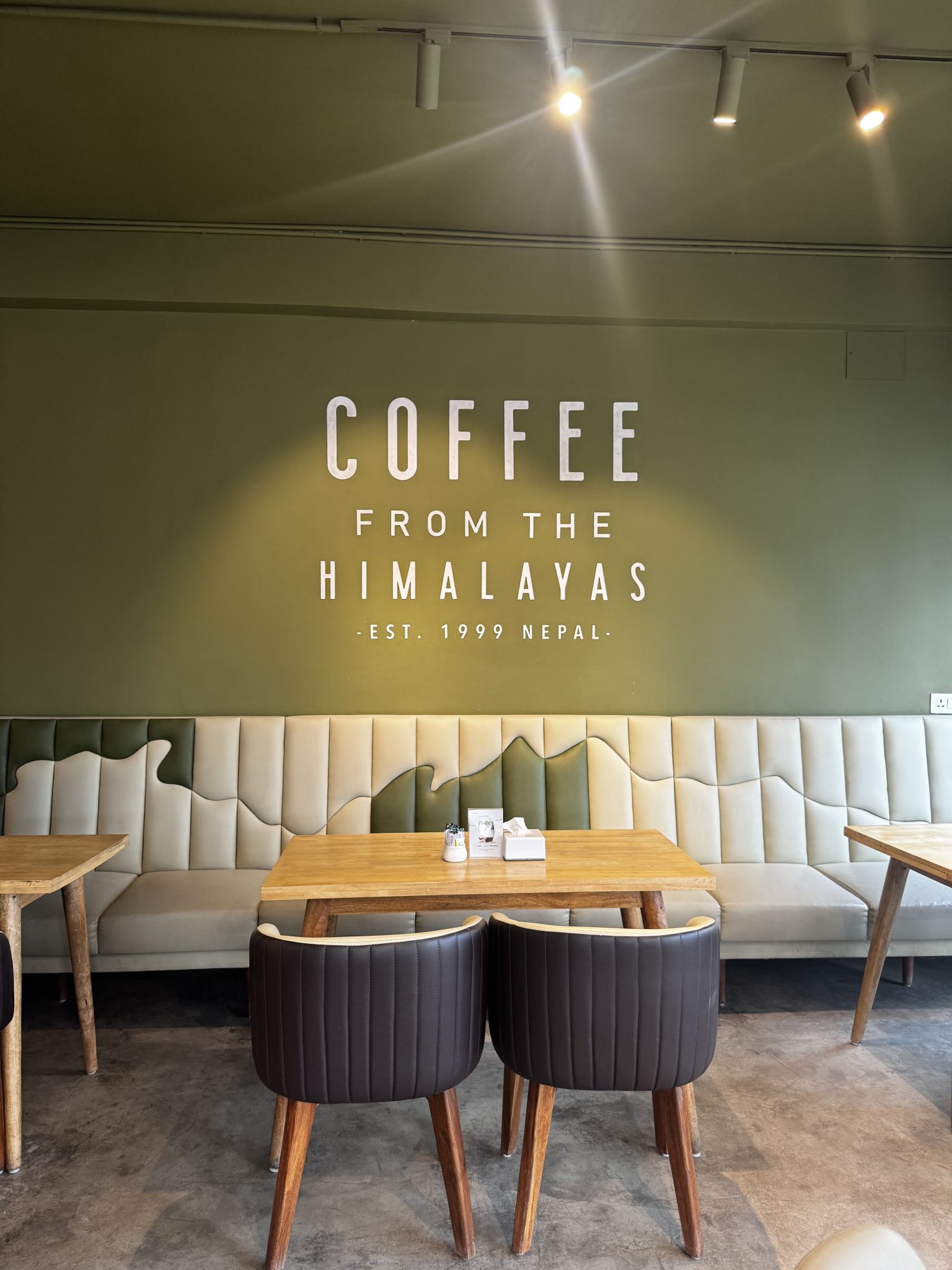 صورة توضيحية (مطاعم ومقاهٍ — A cozy cafe interior with an olive-green wall reading “COFFEE FROM THE HIMALAYAS • EST. 1999 NEPAL.” A light bench with a mountain design lines the wall, with wooden tables and brown chairs in front.) — ليست صورة فعلية لـ ستوريا
